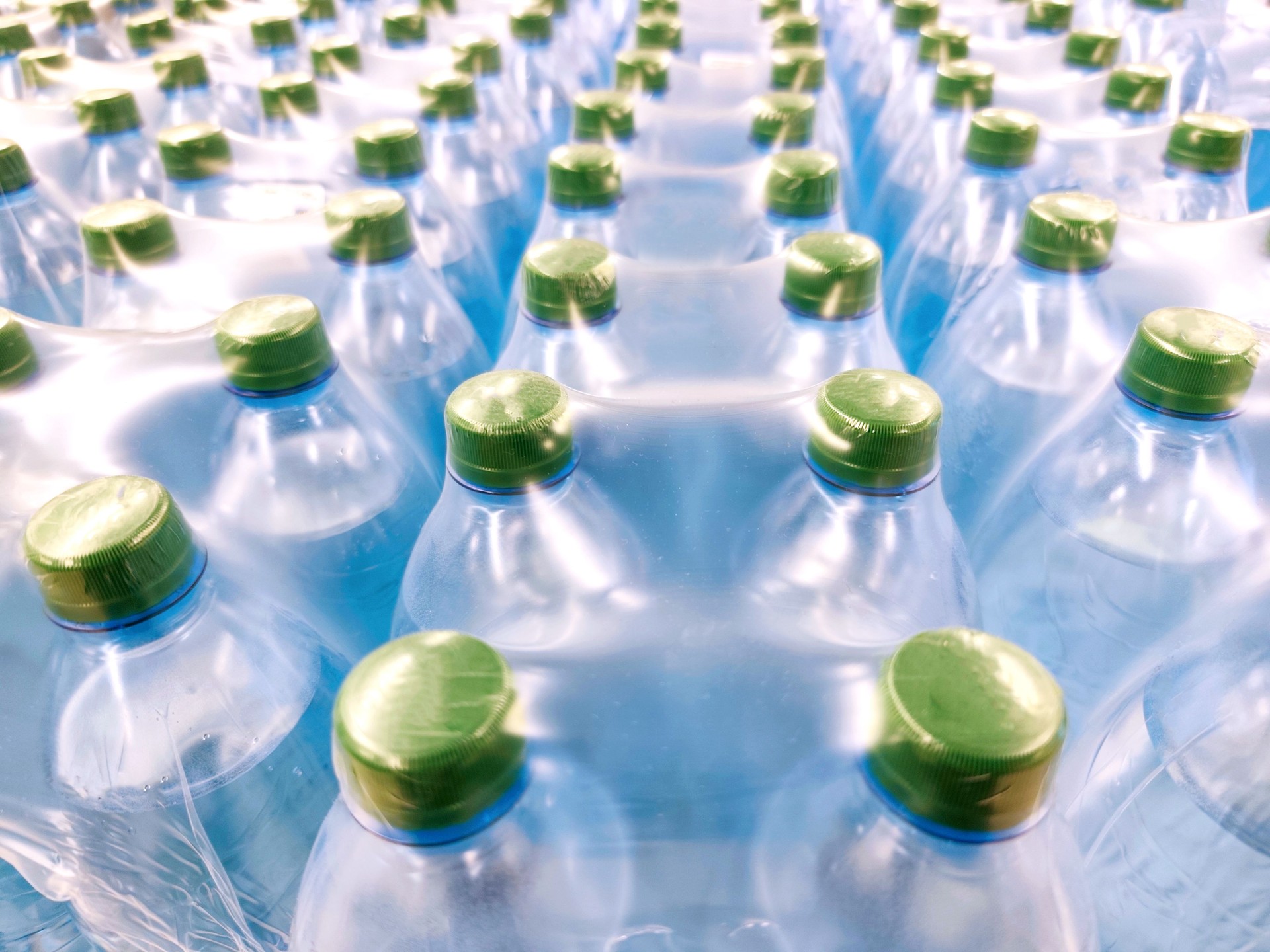 Multiple plastic water bottles tightly packed in shrink wrap, seen from above. Green caps, blue-toned bottles, clean, fresh visual, hydration, mass production, bottled water industry, packaging, and logistics.
